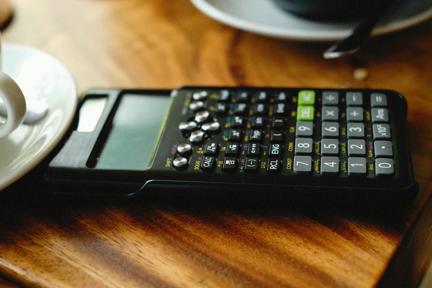 Scientific calculator sitting on a wooden table beside a coffee cup.